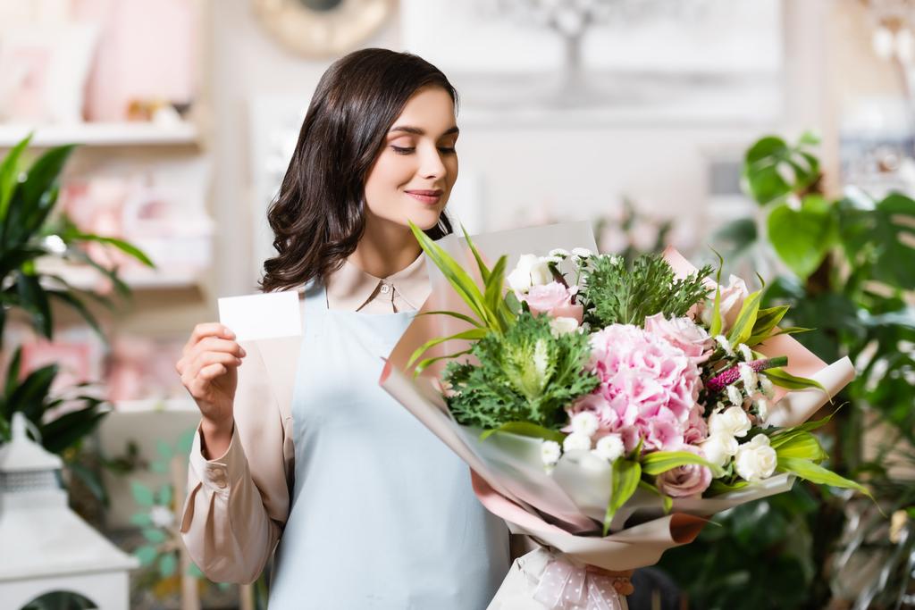 jovem florista olhando para buquê festivo enquanto segurando cartão de visita em branco no fundo borrado - Foto, Imagem