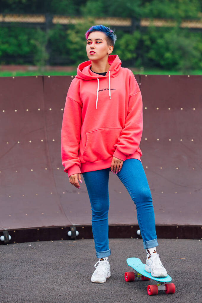 Πορτρέτο ενός trendy όμορφη νεαρή κοπέλα ντυμένη με ροζ κουκούλα και τζιν στέκεται δίπλα στο γήπεδο skateboard με μπλε πλαστικό skateboard της. - Φωτογραφία, εικόνα