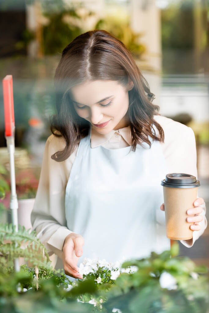 florista sorrindo segurando café para ir ao tocar flores em primeiro plano borrado - Foto, Imagem