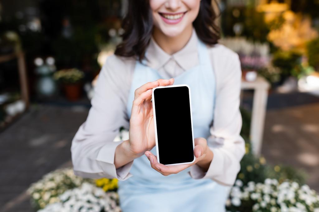 vista recortada de florista sonriente sosteniendo teléfono inteligente con pantalla en blanco cerca de flores en primer plano borroso - Foto, Imagen