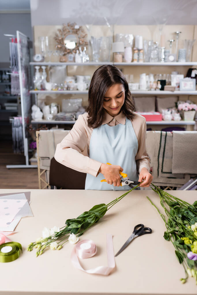 smiling florist cutting steam while making eustoma flowers bouquet near rack with vases on blurred background - Photo, Image
