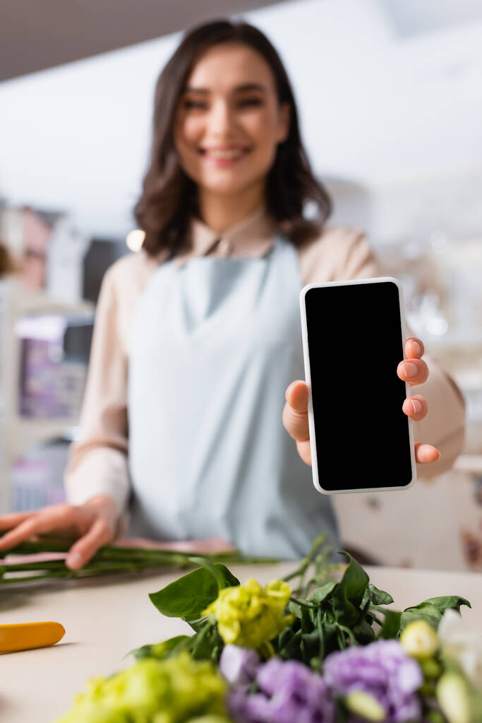 Vista de cerca del teléfono inteligente en la mano de florista sonriente de pie cerca del escritorio con flores de eustoma sobre un fondo borroso - Foto, Imagen