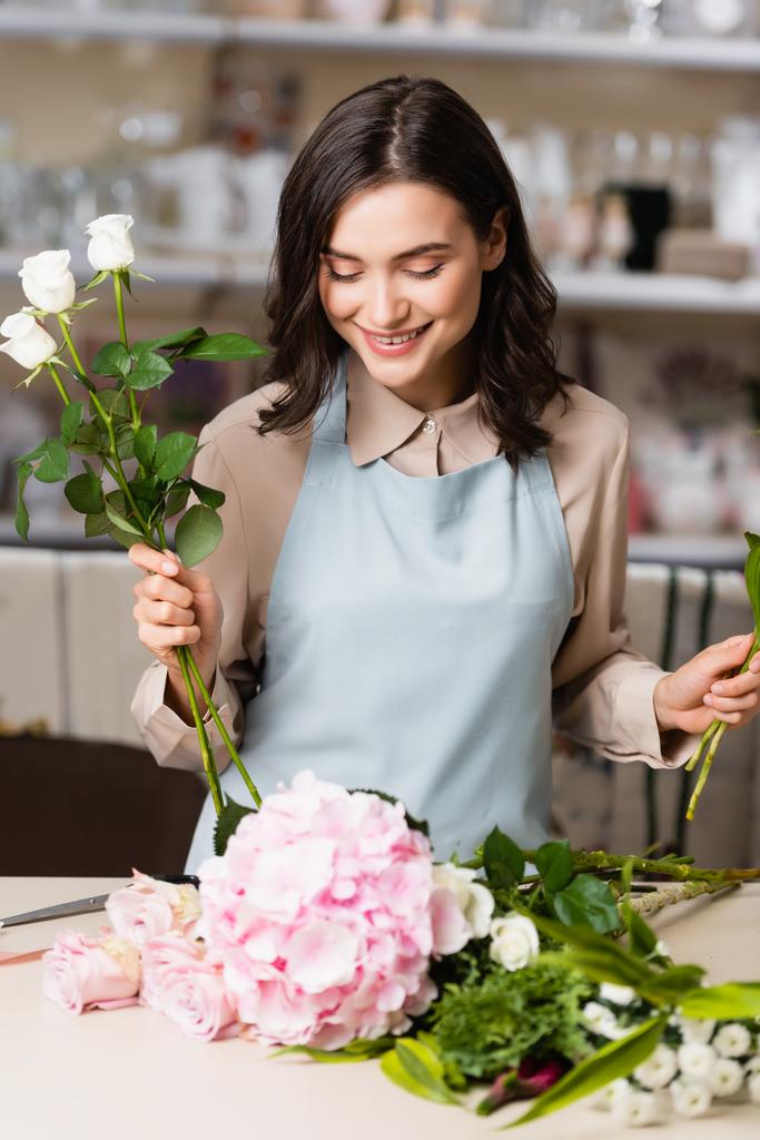 Gelukkige vrouwelijke bloemist met rozen kijken naar bloemen op het bureau, terwijl het componeren van boeket op wazig achtergrond - Foto, afbeelding