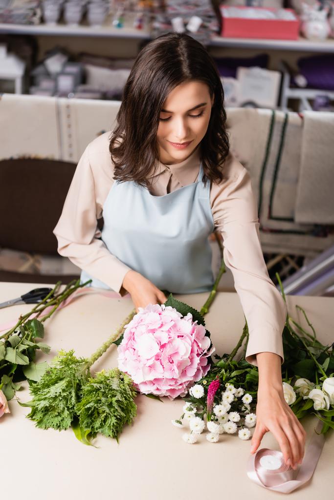 Çiçekçinin çiçekli arka plandaki çiçeklerin yakınındaki masadan dekoratif kurdele aldığı yüksek açılı manzara - Fotoğraf, Görsel