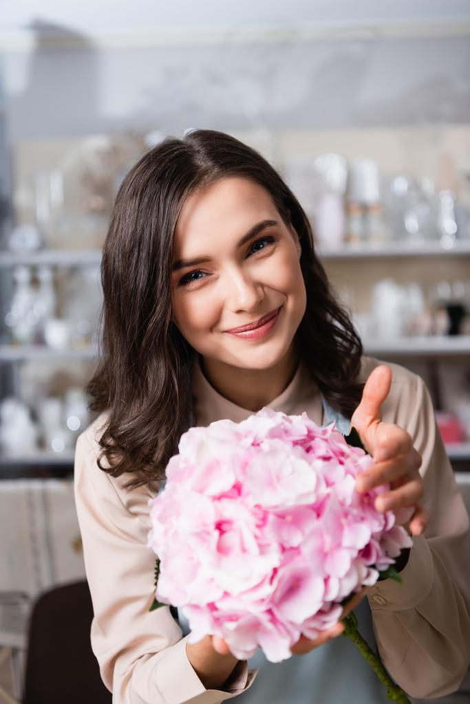 Portrait of happy brunette florist with blooming hydrangea looking at camera on blurred background - 写真・画像