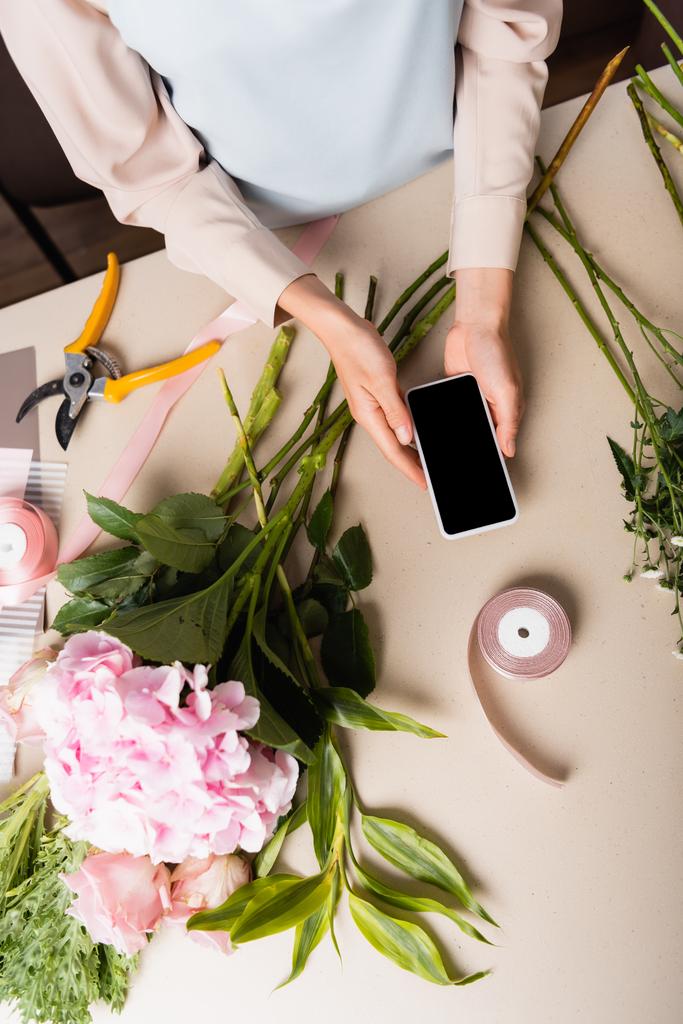 机の上の花の近くに空白の画面でスマートフォンを保持している花屋の作物ビュー - 写真・画像