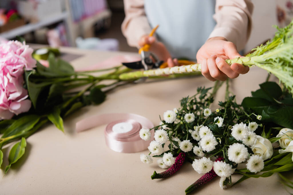 Vista ritagliata del fiorista con forbici fusto di taglio della pianta vicino ai fiori e nastro decorativo sulla scrivania su sfondo sfocato - Foto, immagini