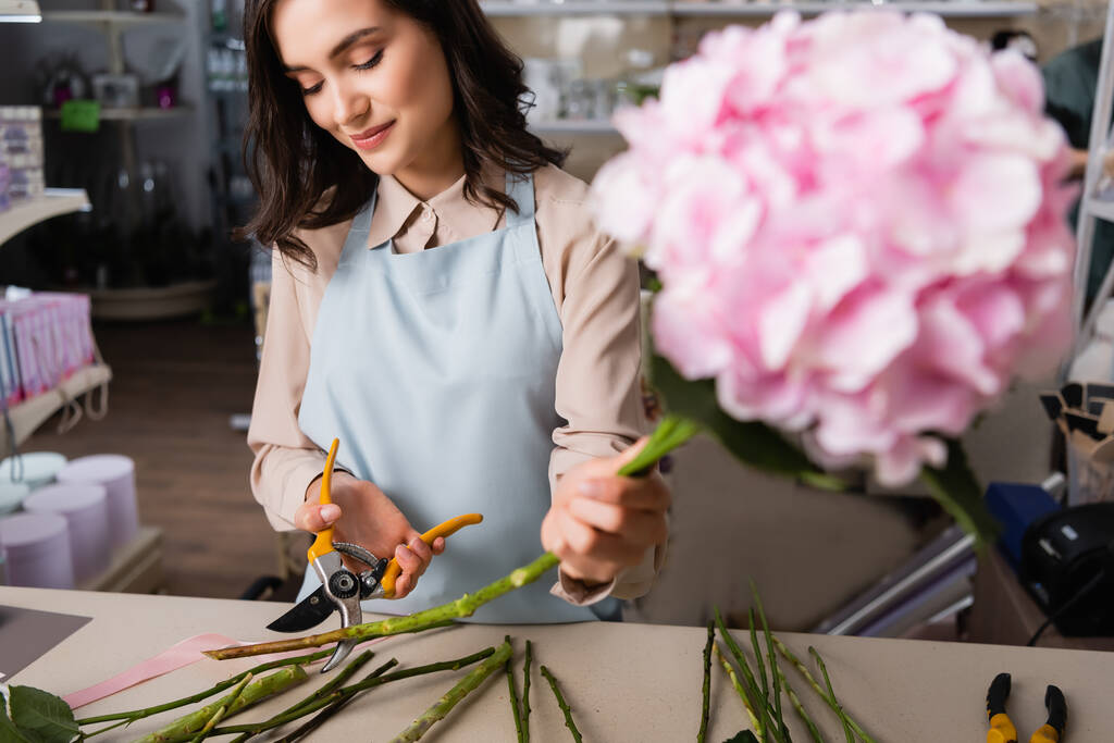 Позитивная женщина-флористка режет стебель растения у стола с размытыми гортензиями на переднем плане - Фото, изображение