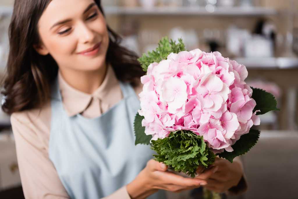 glimlachende vrouwelijke bloemist kijken naar bloeiende hortensia in bloemenwinkel op wazige achtergrond - Foto, afbeelding