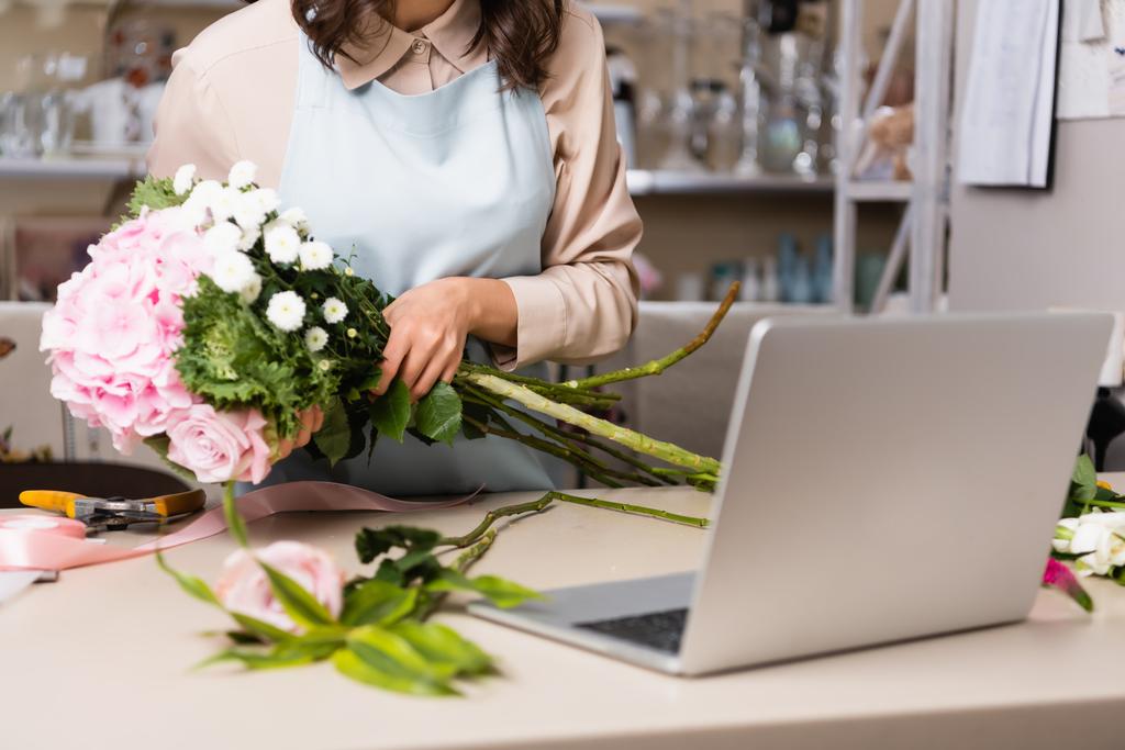 Vista recortada de floristería que arregla ramo con rosas, crisantemos y hortensias cerca de la computadora portátil en el escritorio sobre fondo borroso - Foto, imagen