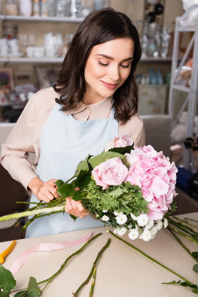 Uśmiechnięta brunetka kwiaciarnia patrząc na bukiet w pobliżu biurka z łodygami na rozmytych regałach na tle - Zdjęcie, obraz