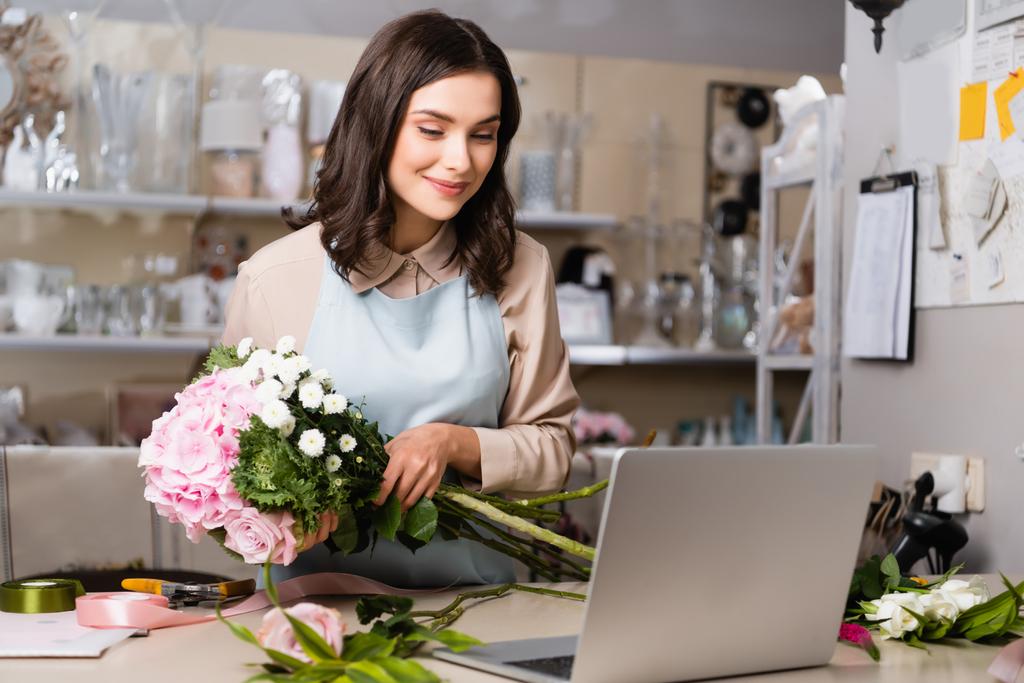 Fleuriste souriante regardant ordinateur portable, tout en arrangeant un bouquet avec des rayonnages flous sur le fond - Photo, image
