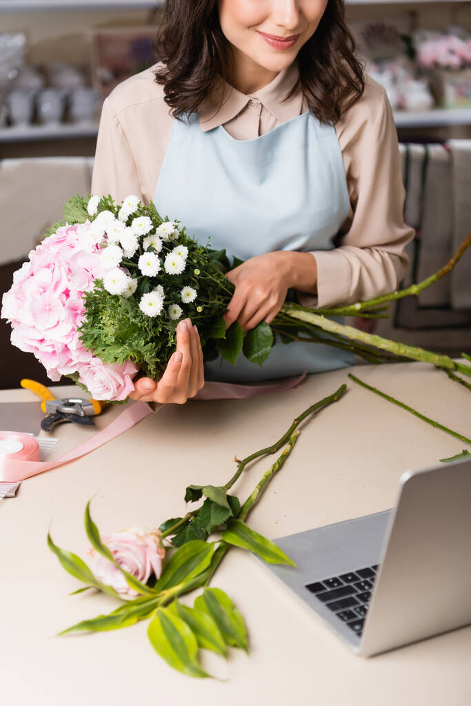 Обрезанный вид флориста, держащего букет с гортензиями, розами и хризантемами рядом с ноутбуком на столе на размытом фоне - Фото, изображение