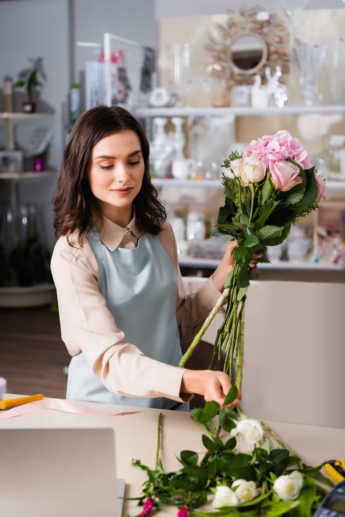 Positivo fiorista femminile prendendo rosa dalla scrivania, mentre organizzare bouquet con rastrelliere sfocate di vasi su sfondo - Foto, immagini