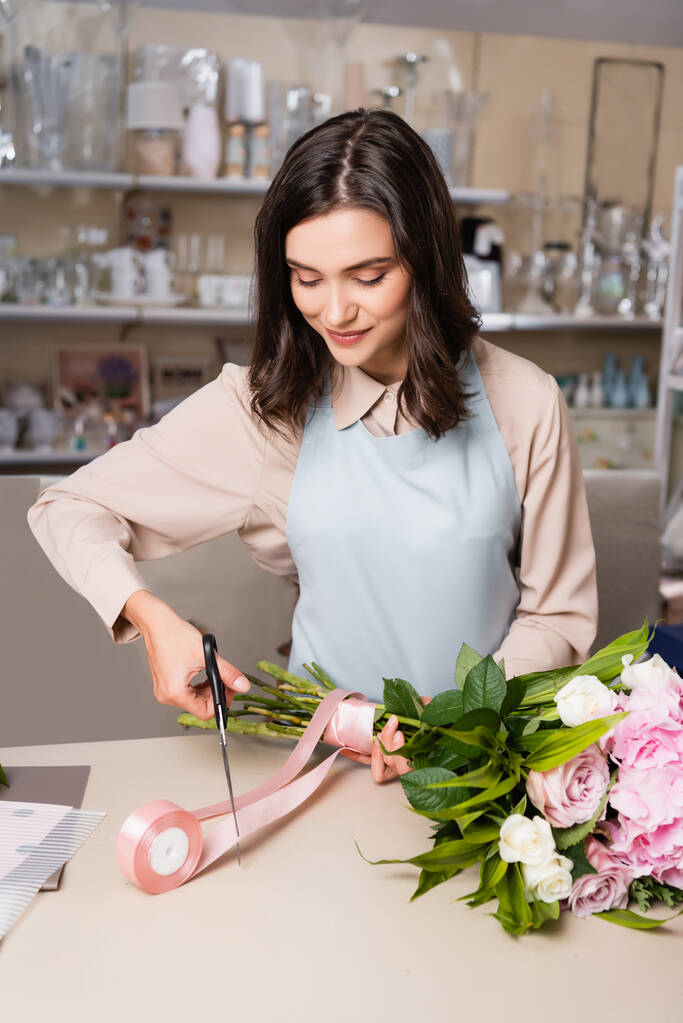 Positive Blumenhändlerin mit Schere, die dekoratives Band schneidet, während sie gebundenen Strauß mit verschwommenen Regalen auf dem Hintergrund hält - Foto, Bild