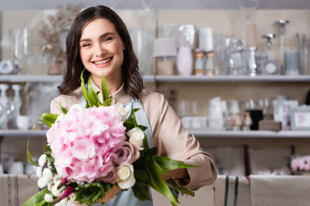 florista feminino feliz olhando para a câmera com buquê florescendo na loja de flores com prateleiras borradas de vasos no fundo - Foto, Imagem
