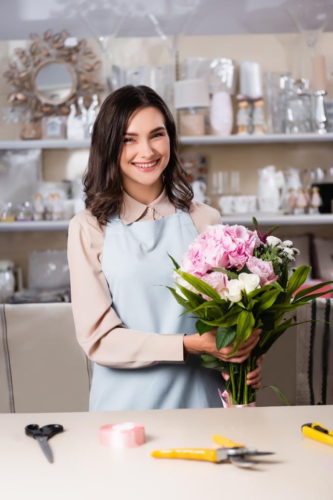 Cheerful florist holding bouquet near desk with tools and decorative ribbon on blurred background - Photo, Image