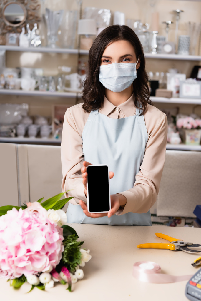 Флорист в медицинской маске показывает смартфон с пустым экраном возле букета на столе с размытыми стеллажами ваз на заднем плане - Фото, изображение