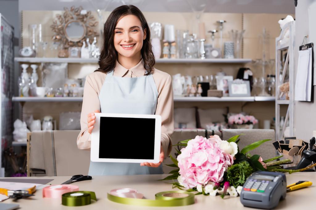 Fiorista felice mostrando tablet digitale vicino bouquet sulla scrivania con rastrelliere sfocate di vasi su sfondo - Foto, immagini