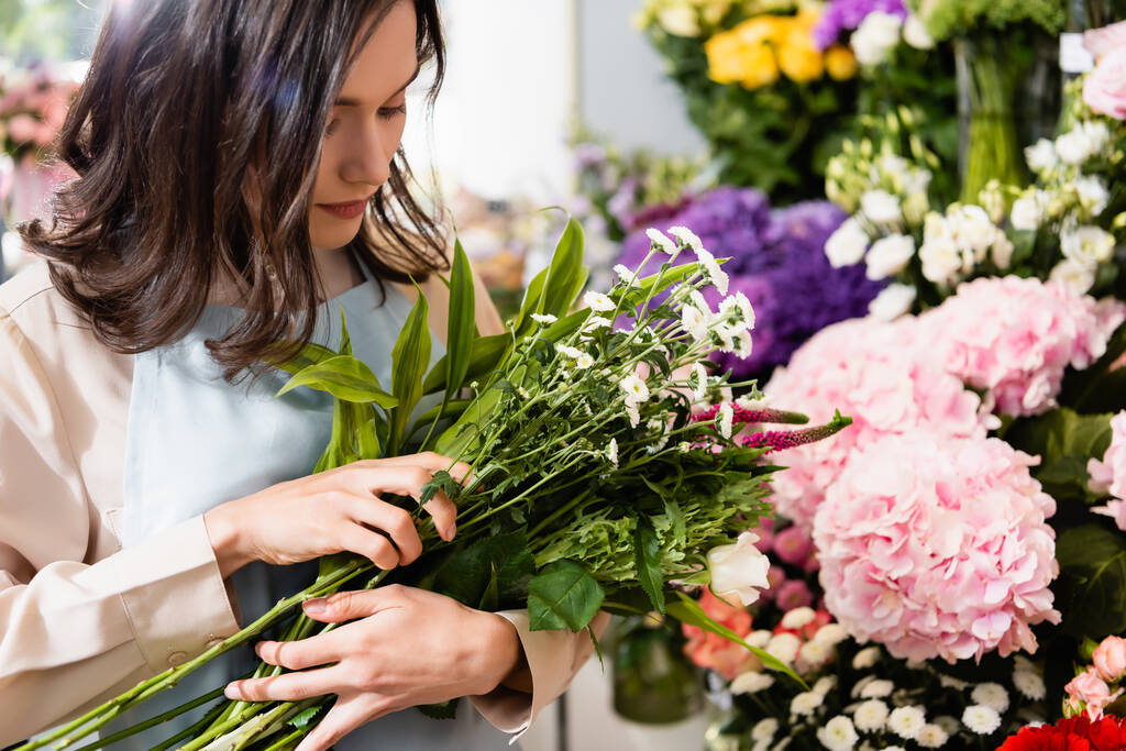 Female florist arranging bouquet with celosia nd chrysanthemums with blurred flowers on background - Photo, Image