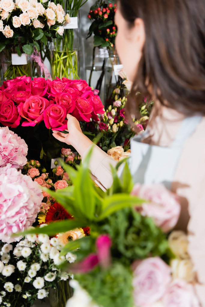 Brunette florist touching rouses in vase on rack of flowers with blurred bouquet on foreground - Photo, Image