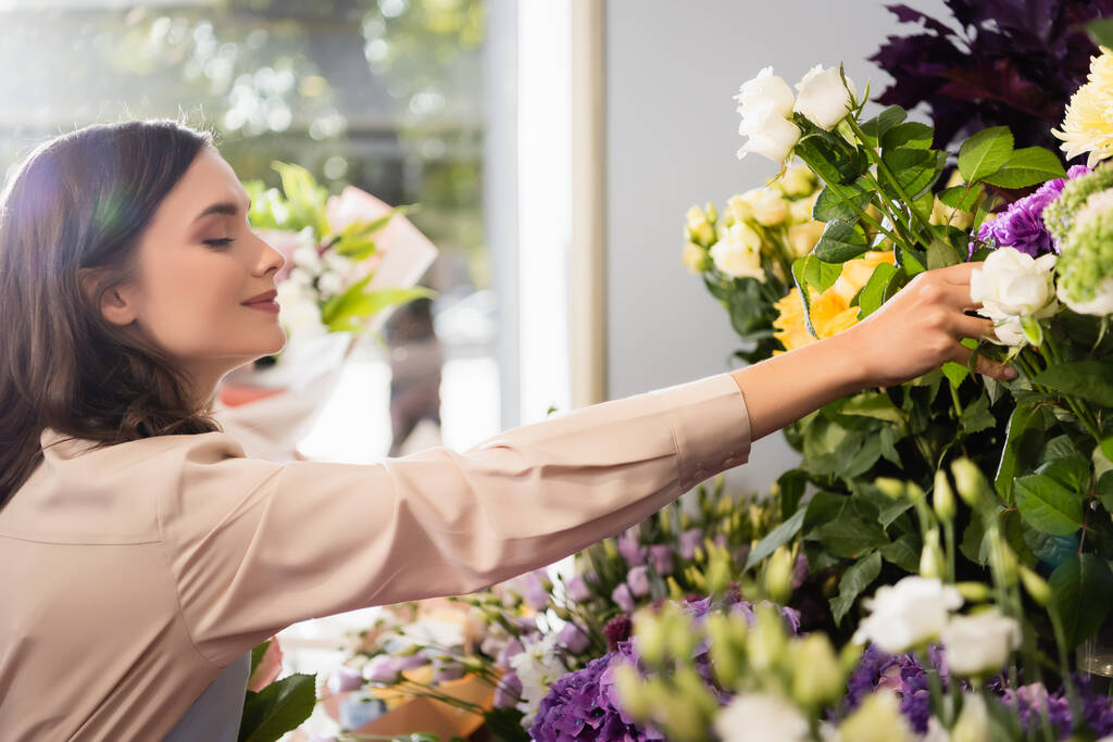 Πλευρική άποψη του ευτυχισμένη γυναίκα ανθοπώλης λαμβάνοντας αυξήθηκε κοντά ράφια των λουλουδιών με θολή παράθυρο σε πρώτο πλάνο - Φωτογραφία, εικόνα