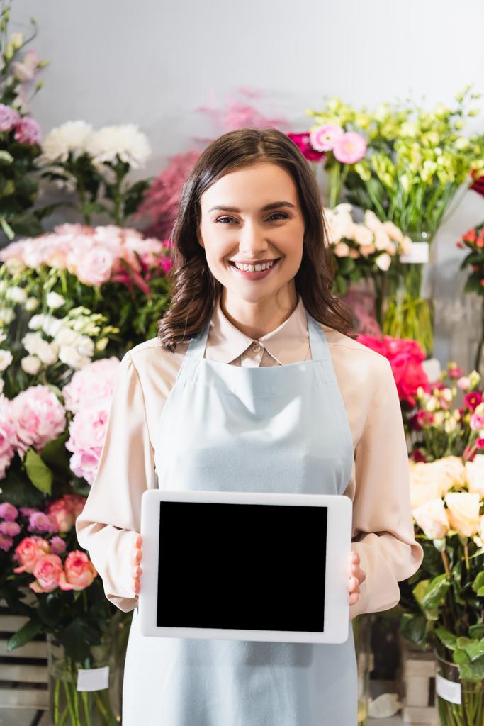 Frontansicht eines glücklichen Blumenhändlers, der in die Kamera schaut, während er ein digitales Tablet mit verschwommenen Blumenständern auf dem Hintergrund hält - Foto, Bild