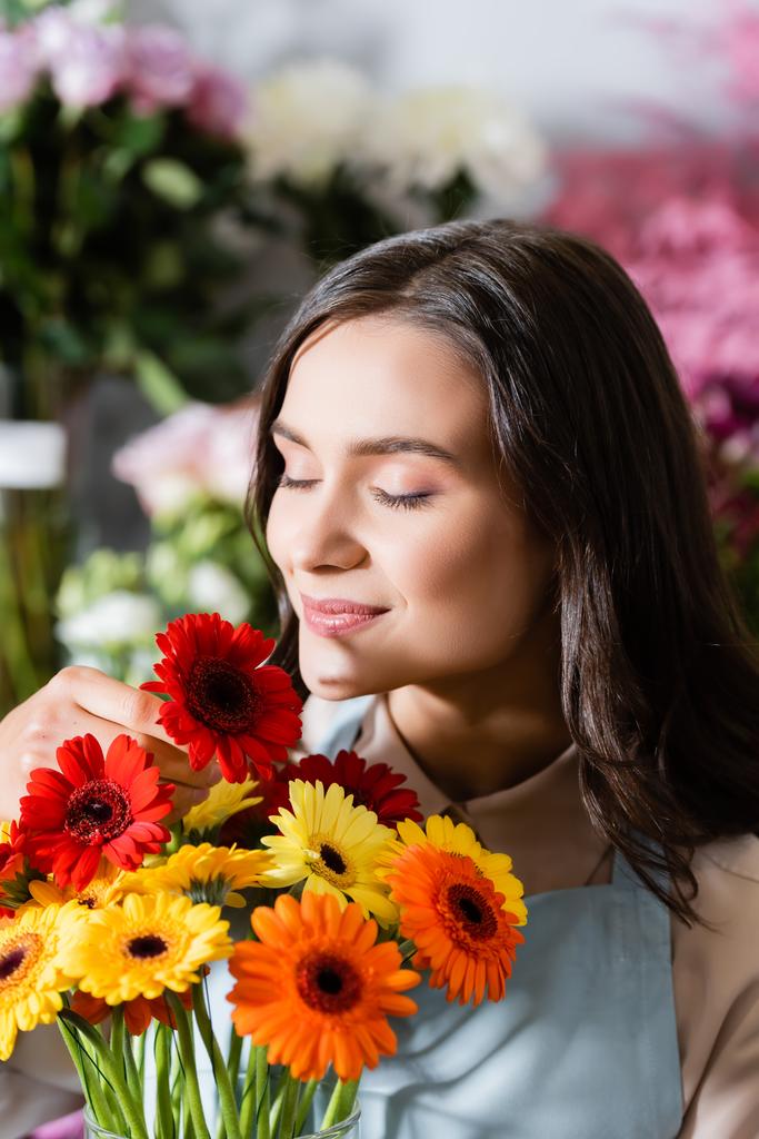 Florista femenina sonriente con ojos cerrados oliendo gerberas con flores borrosas en el fondo - Foto, imagen
