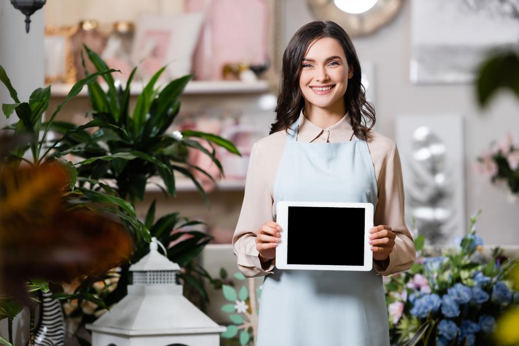 Счастливая флористка смотрит в камеру, держа в руках цифровой планшет в цветочном магазине на размытом переднем плане - Фото, изображение