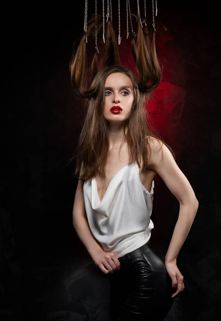 Ένα όμορφο κορίτσι με βραδινό μακιγιάζ και κόκκινα χείλη που φοράει μπλούζα με βαθύ ντεκολτέ και τα μαλλιά της κρεμασμένα στις αλυσίδες, ποζάροντας σε κόκκινο και μαύρο φόντο. Εννοιολογικός, πρωτοποριακός σχεδιασμός. - Φωτογραφία, εικόνα