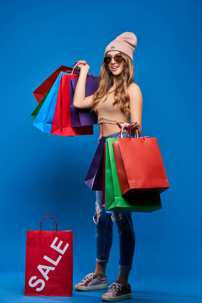 Beautiful young woman in sunglasses and a hat with packages on sale. Happy girl after shopping on blue background. - Photo, Image
