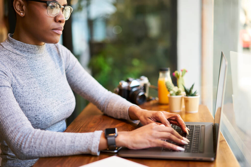 Seitenansicht einer konzentrierten schwarzen Frau, die am Computer im Internet surft, während sie im modernen Café an einem Finanzbericht arbeitet - Foto, Bild