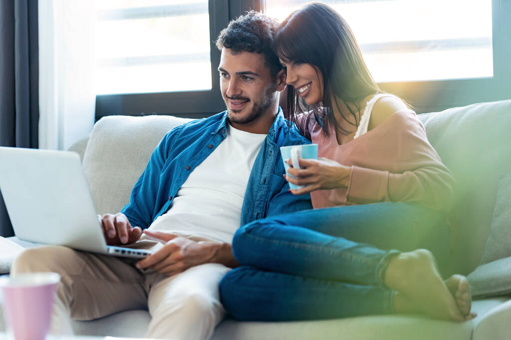 Tiro de feliz hermosa pareja usando el ordenador mientras está sentado en el sofá en casa. - Foto, imagen