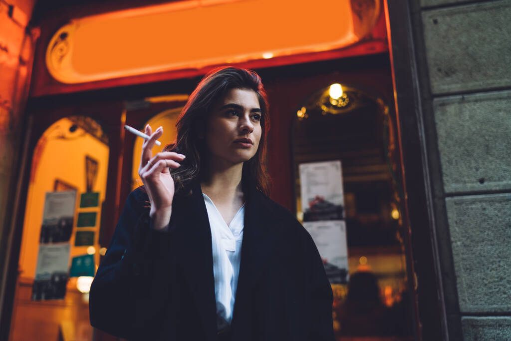 De baixo de sério feminino pensativo em desgaste formal de pé na rua e fumar cigarro enquanto olha para longe durante a caminhada - Foto, Imagem