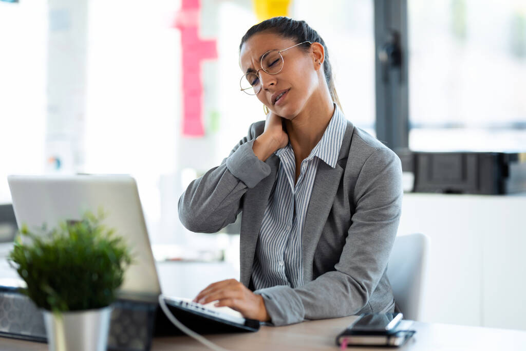 Egy fáradt, nyaki fájdalommal küszködő fiatal nő, aki az irodában dolgozik a számítógépével. - Fotó, kép