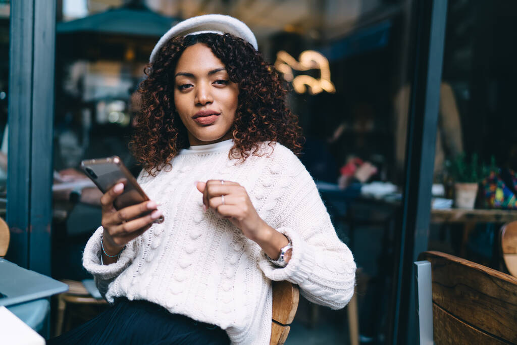 Ethnische junge Frau in weißer Baskenmütze und in lässiger Kleidung sitzt am Tisch und tippt mit dem Handy SMS, während sie sich in einem gemütlichen Café entspannen und in die Kamera schauen - Foto, Bild