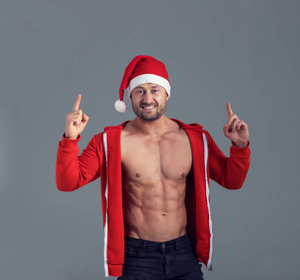 Giovane uomo muscoloso in costume da Babbo Natale punta le dita verso l'alto mentre in piedi su uno sfondo grigio. - Foto, immagini