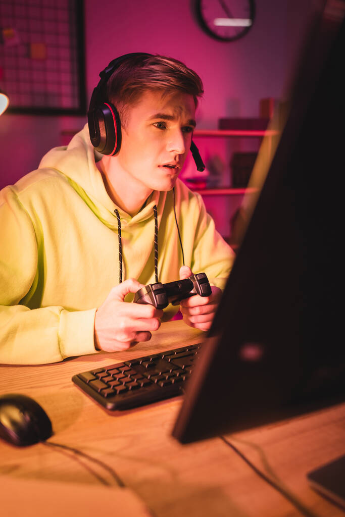 KYIV, UKRAINE - AUGUST 21, 2020: Young man in headset playing video game with joystick and computer on blurred foreground  - Photo, Image