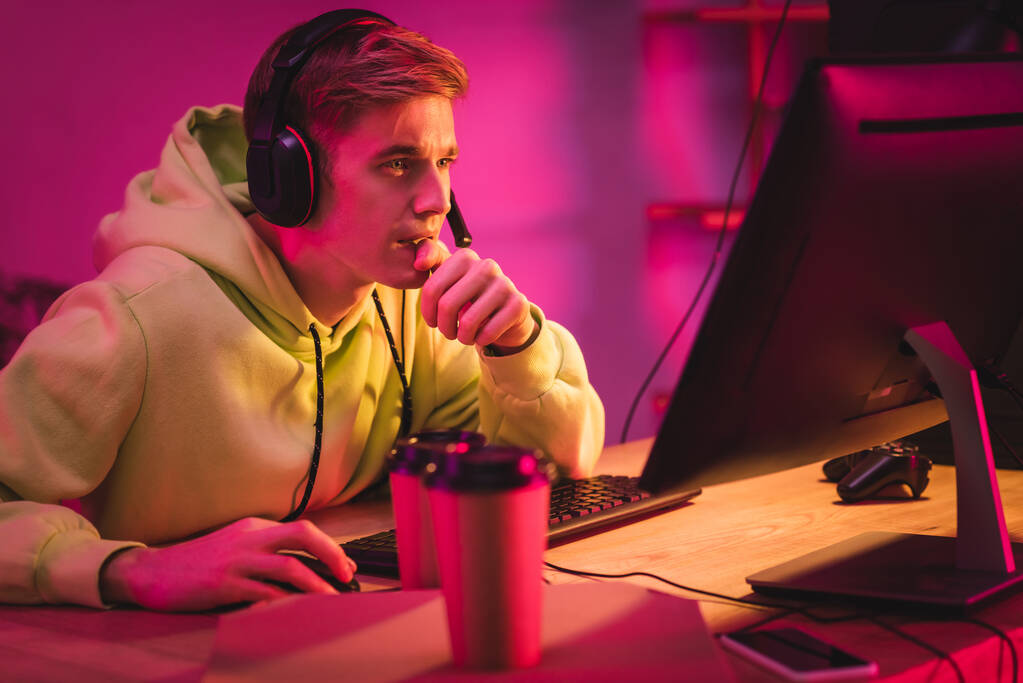KIEW, UKRAINE - 21. AUGUST 2020: Fokussierter Gamer im Headset mit Computer in der Nähe von Coffee to go, Pizzakiste und Steuerknüppel im verschwommenen Vordergrund  - Foto, Bild