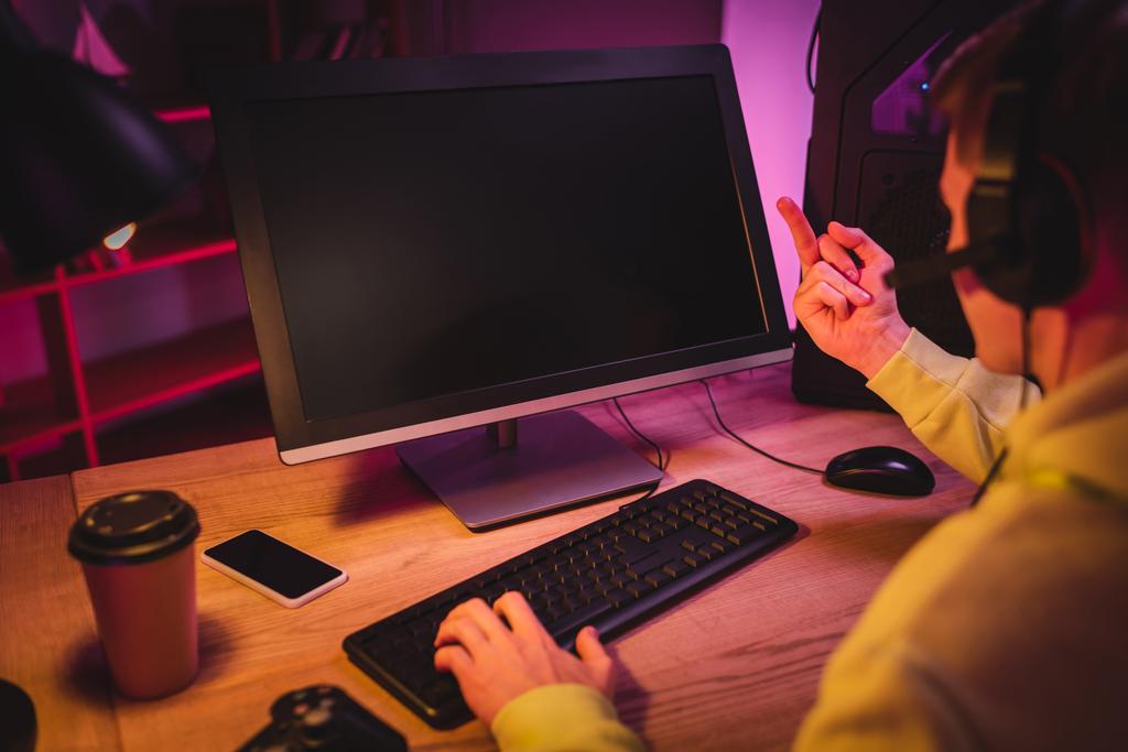 Плеер в гарнитуре, показывающий средний палец на мониторе компьютера, играя в видеоигру рядом со смартфоном и отнимая кофе на размытом переднем плане  - Фото, изображение