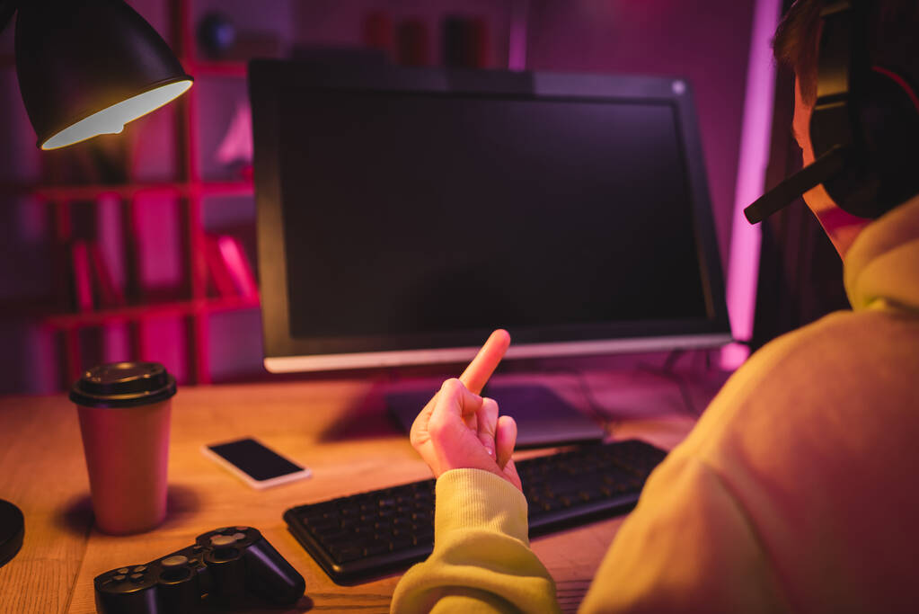 KYIV, UCRANIA - 21 de agosto de 2020: Gamer en auriculares que muestran el dedo medio cerca del joystick, café para llevar y computadora sobre fondo borroso  - Foto, imagen