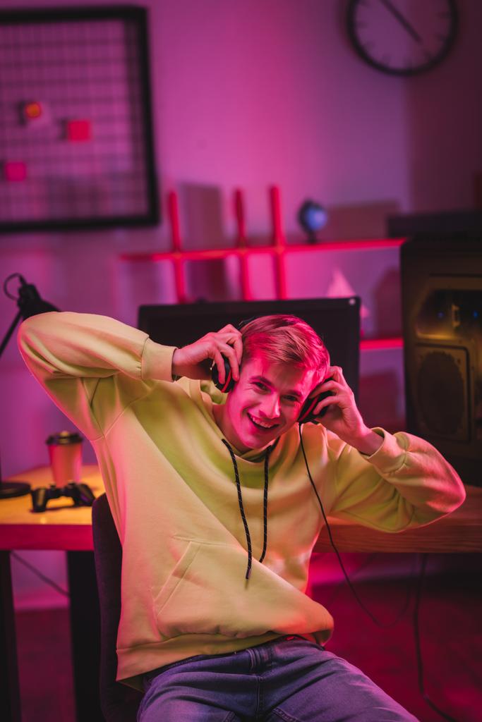 Веселый геймер держит наушники рядом с компьютером и джойстиком на размытом фоне  - Фото, изображение