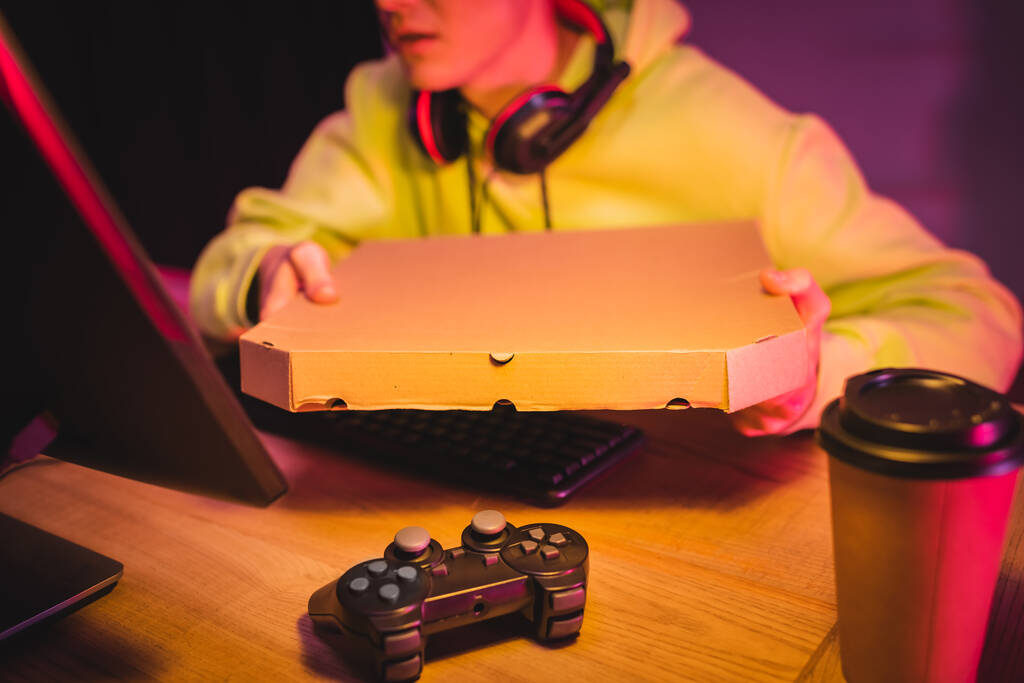 KYIV, UKRAINE - AUGUST 21, 2020: Joystick and coffee to go near gamer in headphones holding pizza box near computer on blurred background  - Photo, Image