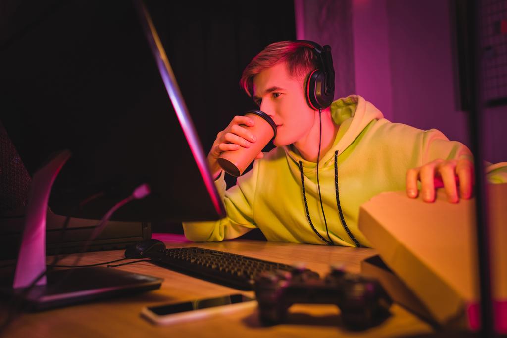 Mladý hráč ve sluchátkách pití kávy jít a držet pizza box v blízkosti počítače a joystick na rozmazané popředí  - Fotografie, Obrázek