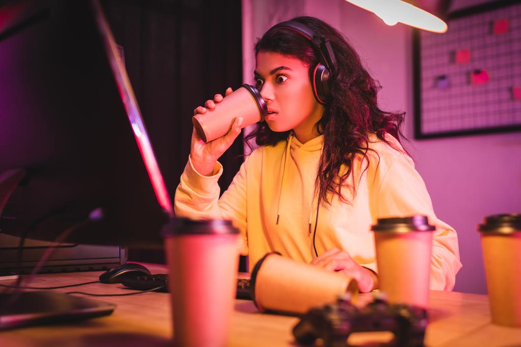 Izgalmas afro-amerikai játékos headset iszik kávét ho közelében számítógép és joystick elmosódott előtérben  - Fotó, kép