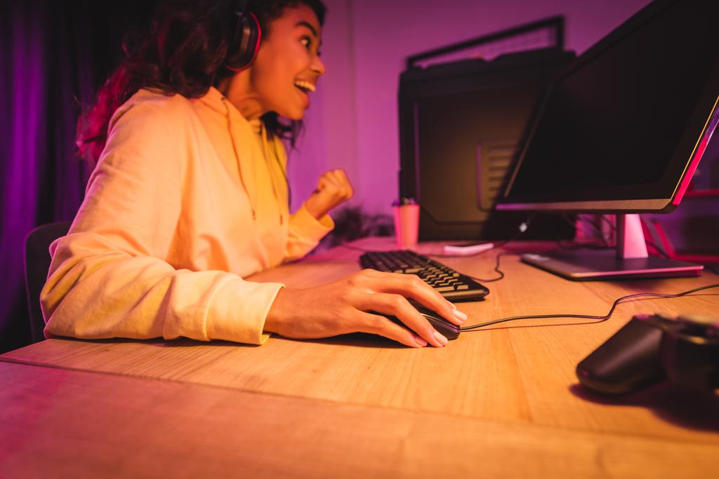 Joueur afro-américain joyeux utilisant l'ordinateur et montrant geste ouais près joystick sur le premier plan flou  - Photo, image