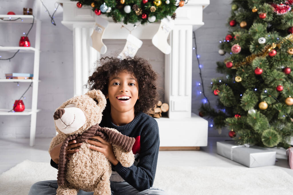 excitada chica afroamericana mirando a la cámara mientras sostiene el oso de peluche cerca de la chimenea decorada y el árbol de Navidad sobre un fondo borroso - Foto, Imagen