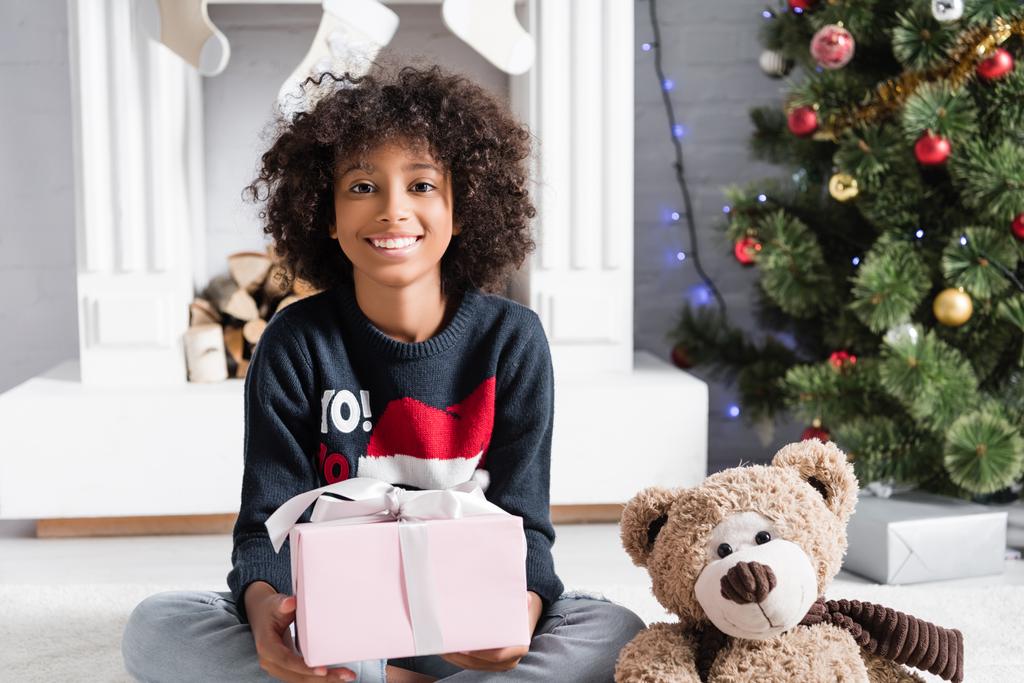 χαρούμενη Αφροαμερικανή κοπέλα που κοιτάζει την κάμερα κρατώντας ένα δώρο κοντά στο χριστουγεννιάτικο δέντρο και το τζάκι σε θολή φόντο - Φωτογραφία, εικόνα