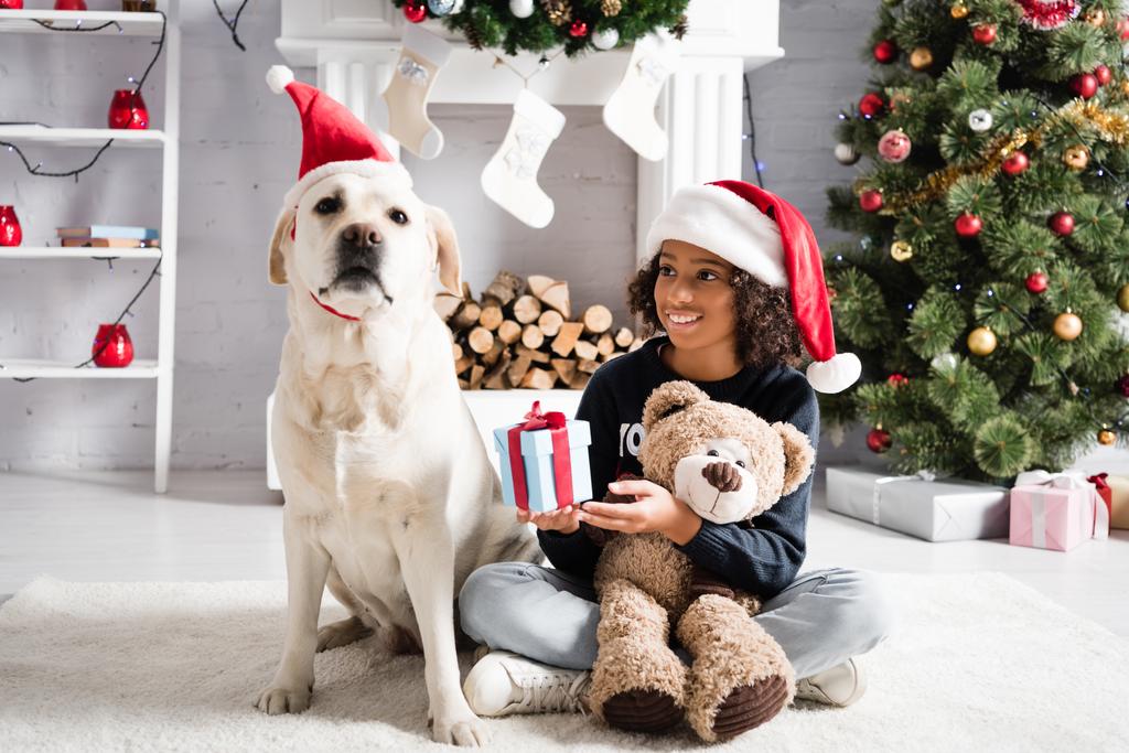feliz afroamericana chica sosteniendo caja de regalo y osito de peluche mientras está sentado en el suelo cerca de perro labrador sobre fondo borroso - Foto, imagen
