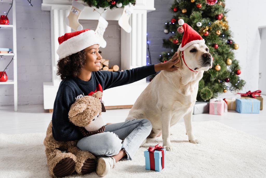 joyful african american girl stroking labrador dog while sitting on floor with teddy bear near gift box on blurred background - Photo, Image